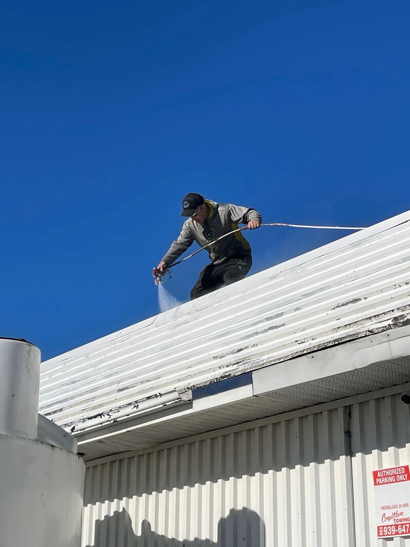 A crew member standing on a metal roof, pressure washing it with a long wand.