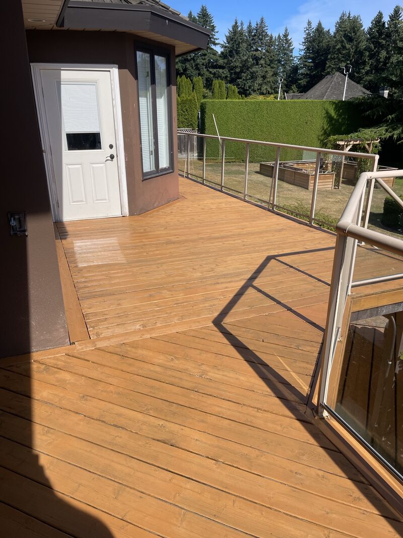 A deck stained a warm wood tone, with a glass railing and a white door.