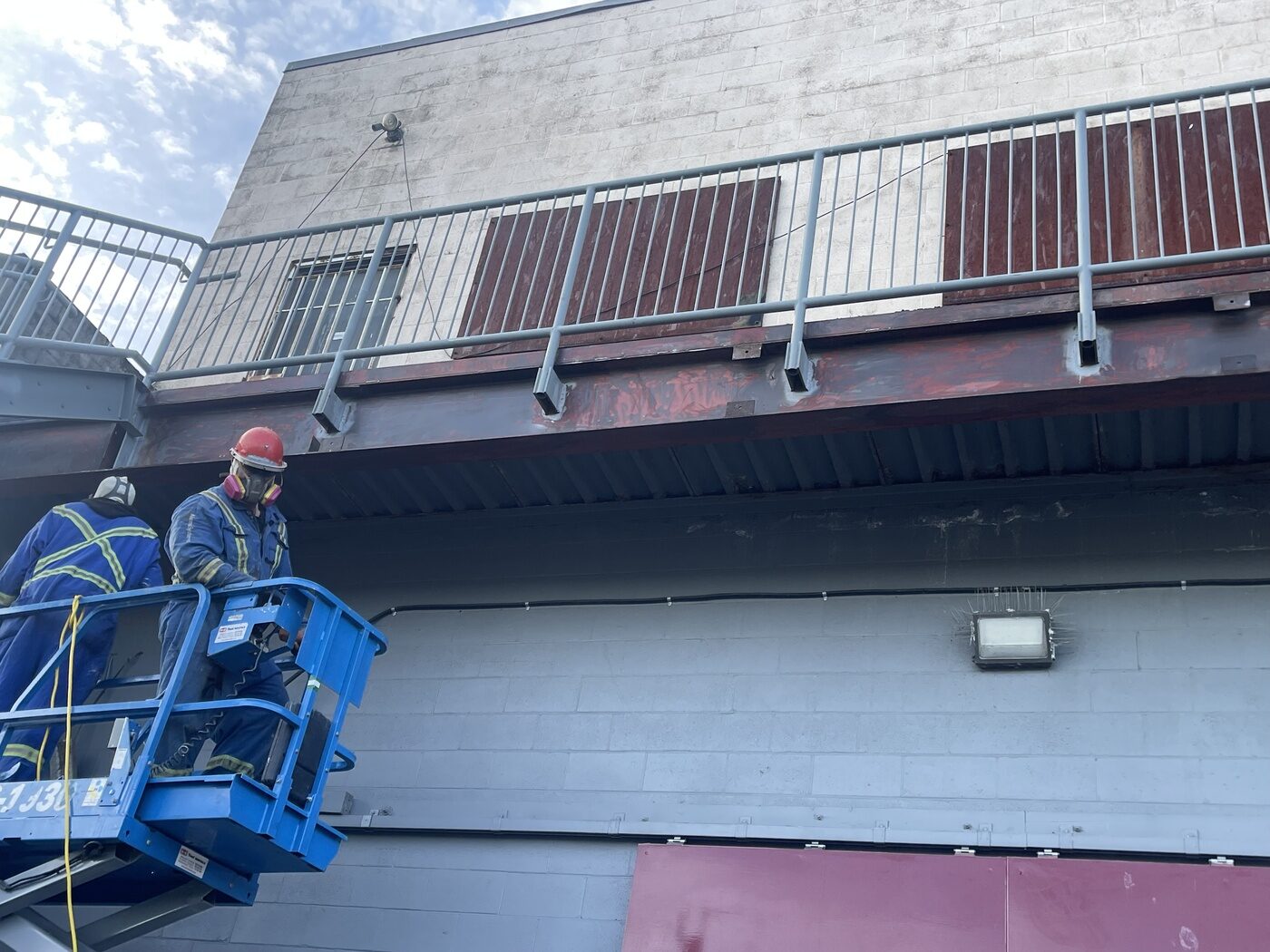 Two painters in coveralls, hard hats and respirators working from a blue scissor lift on a rusted steel beam beneath a walkway.