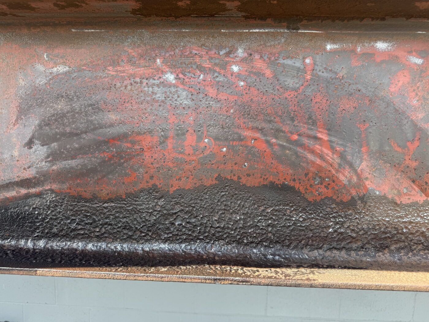 Close-up of a structural steel beam: corroded, pitted surface with remnants of red primer above, and a band of clean, dark, abrasive-blasted steel below.